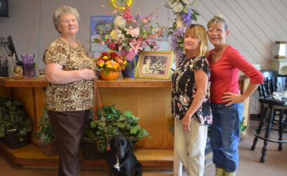 Bobbie McElhaney with Sadie (left), Raye Lyne McElhaney, and Jenniffer Miller with a display of arrangements and a photo of the late Gertrude Tubbs Baugh, founder of Tubbs of Flowers, where they all work. Bobbie McElhaney is president of the company, which was started by Baugh in 1946. Photo by Jane Kathleen Gregorio