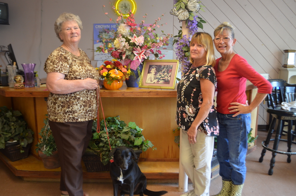 Bobbie McElhaney with Sadie (left), Raye Lyne McElhaney, and Jenniffer Miller with a display of arrangements and a photo of the late Gertrude Tubbs Baugh, founder of Tubbs of Flowers, where they all work. Bobbie McElhaney is president of the company, which was started by Baugh in 1946. Photo by Jane Kathleen Gregorio